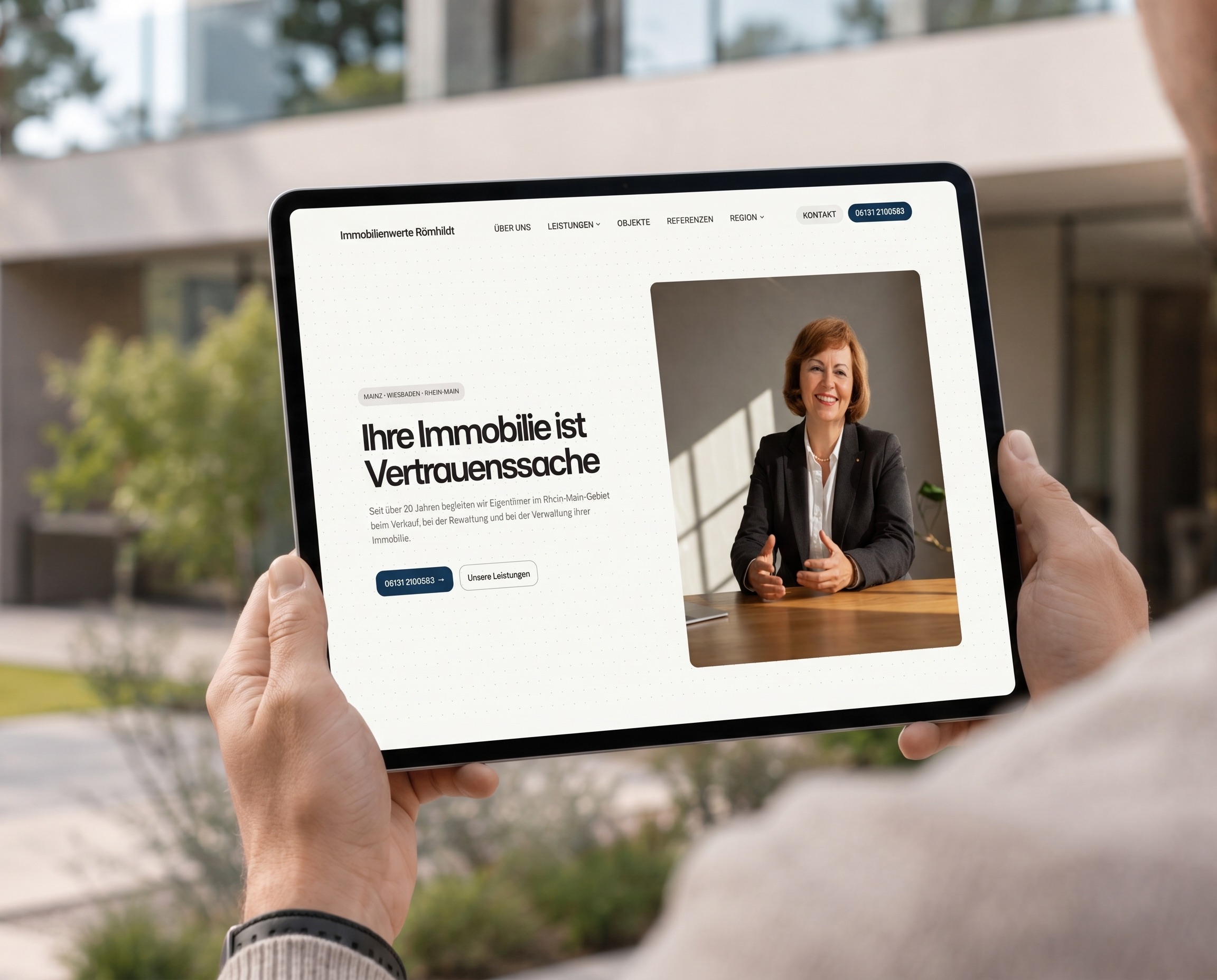 Startseite von Immobilienwerte Römhildt auf einem Tablet, gehalten vor einem modernen Haus mit Garten: „Ihre Immobilie ist Vertrauenssache“ neben dem Foto einer Frau im Blazer am Besprechungstisch